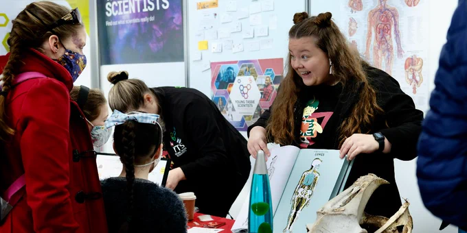 People at a science fair. One person is holding open a book that that contains a picture of the human skeleton and is explaining the image to two children who are accompanied by an adult