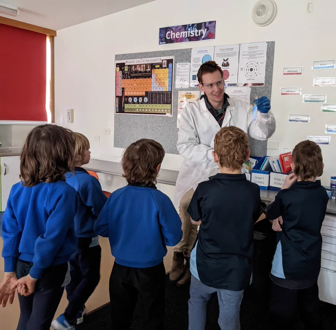 A person standing in a classroom wearing a lab coat and holding a beaker, they are surrounded by a group of children 