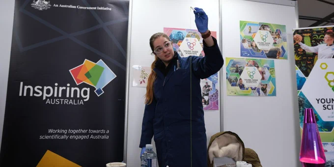 A person dressed in laboratory clothes including goggles, gloves and a gown. They are doing a science experiment that involves pulling a stretchy yellow object out of a beaker with a pair of tweezers 