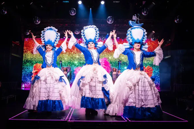 Three cast members wear big blue and white dresses and wigs as they hold hands in the air