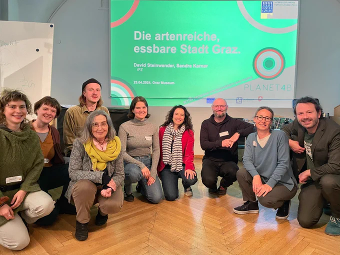 A group of 9 people sitting on their haunches in front of a green projected slide on the case of the biodiverse edible city of Graz.