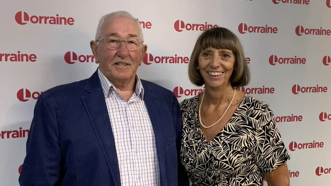 Derek is on the left and is dressed in a navy jacket and checked white shirt. Jean is on the left and is in a white and black leaf-patterned dress. 
They are pictured in front of a screen with the words Lorraine in pink.