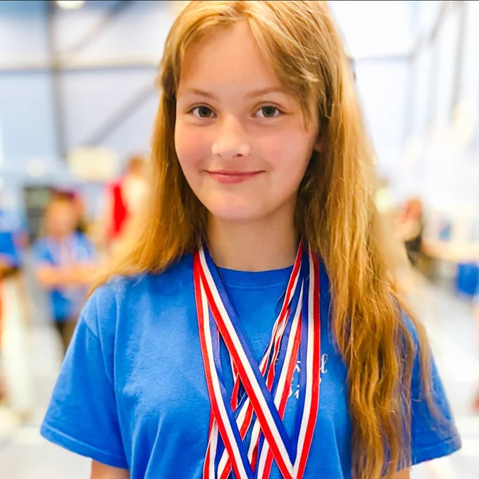 Aaliyah stands in her school sports kit, she is smiling and wearing multiple medals. She has long hair and the background is blurred. 