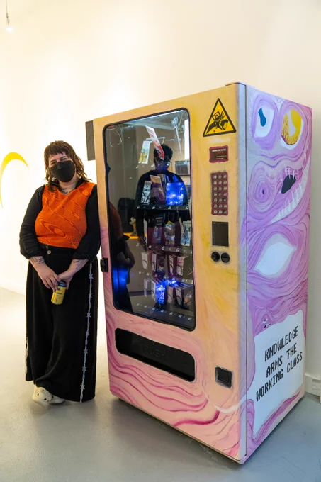 A photo of Sid Drmay standing next to the Zine Machine in the Hamilton Artists Inc James Gallery. Sid is a white person with medium brown hair. They're wearing a black skirt, an orange shirt with long black sleeves and a black KN95 mask. They smile proudly at the camera.