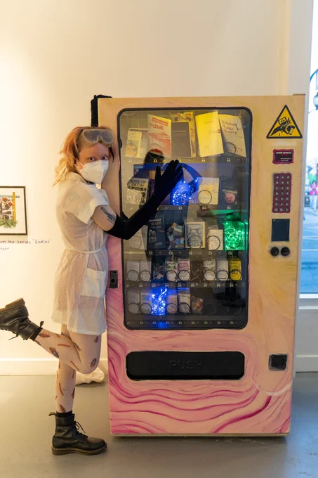 A photo of Venus Underhill, curator and artist, standing next to the zine machine. Venus is a white person with medium length hair. He is wearing a white dress and long black arm gloves, the right hand of which is resting on the glass of the zine machine. They are also wearing eye protection on their forehead and a white KN95 mask.