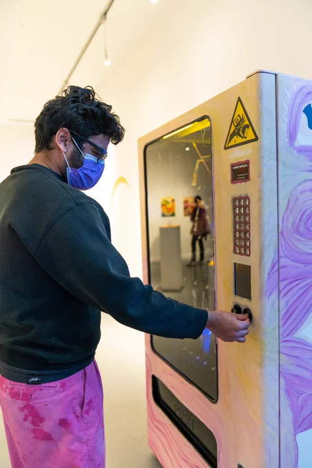 A photo of Nikhil, a person with dark hair and brown skin, inserting a token into the zine machine. Nikhil is wearing a purple mask, a green sweaater, and pink pants with red abstract patterns.