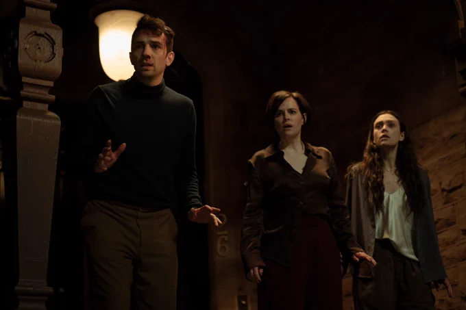 A film still from Humane. A man and two women stand next to each other with concerned expressions outside. The man wears a black long-sleeve shirt and his head is bleeding.