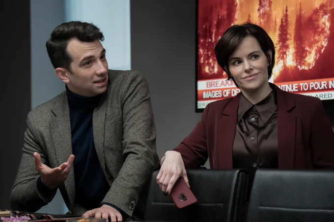 A film still from Humane. A man and woman sit next to each other, they both wear suits and have small smiles on their faces. Behind them is a TV screen showing a news segment of trees on fire.