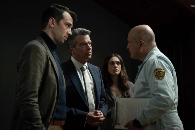 A film still from Humane. Two men in suits stand in front of a man in a uniform holding a clipboard. They all have serious expressions. Behind them is a woman with long brown hair who also stares at the man in uniform with a serious expression.