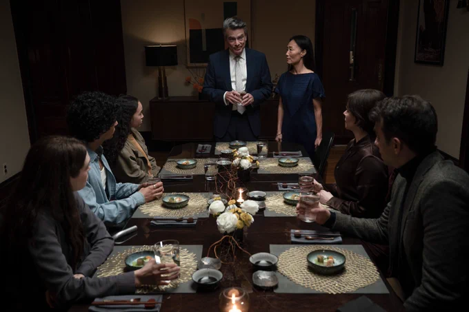 A film still from Humane. A group of people sit together at a dining table with the same dish and placemats in front of them. Two of them are standing, a man and a woman. The man wears a suit, holding a drink, and stares at the group sitting down while the woman standing next to him looks up at him with a serious expression.