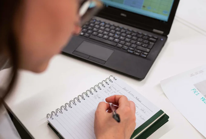 A female researcher is making notes while working on a project in front of her laptop