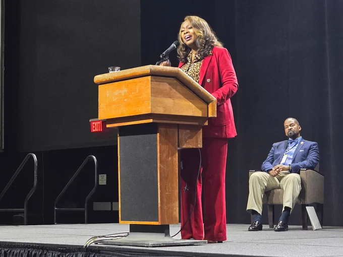 A woman in a red suit speaks at a podium during a conference, while a man in a suit sits behind her on stage, listening attentively.