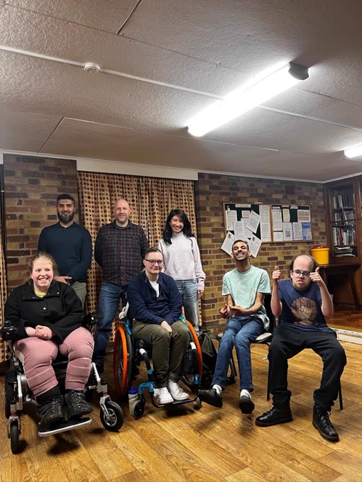 The Ofcom team (back row) with (from left to right) Joanna, Joey, Prash and Thomas. Joanna and Joey are using wheelchairs, Prash and Thomas are seated and Thomas is giving two thumbs up to the camera. They are inside a community venue which has brick walls, a wooden floor and noticeboards. Everyone is smiling.