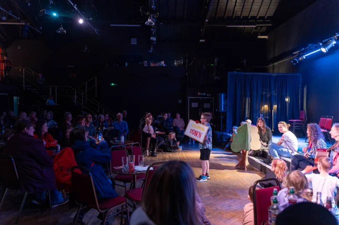 A young boy holds up a sign that reads Kindness Town as part of a performance in our Made In Millom event. The audience look on with smiles on their faces. 
