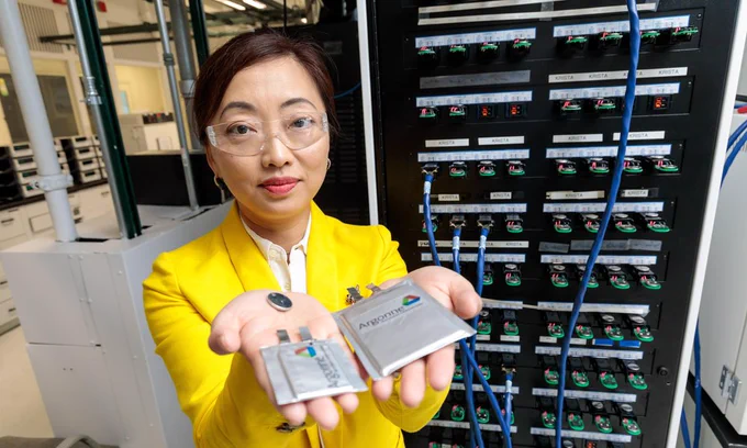 A photograph of a woman wearing safety goggle and a yellow suit. She is holding three batteries in her hands.