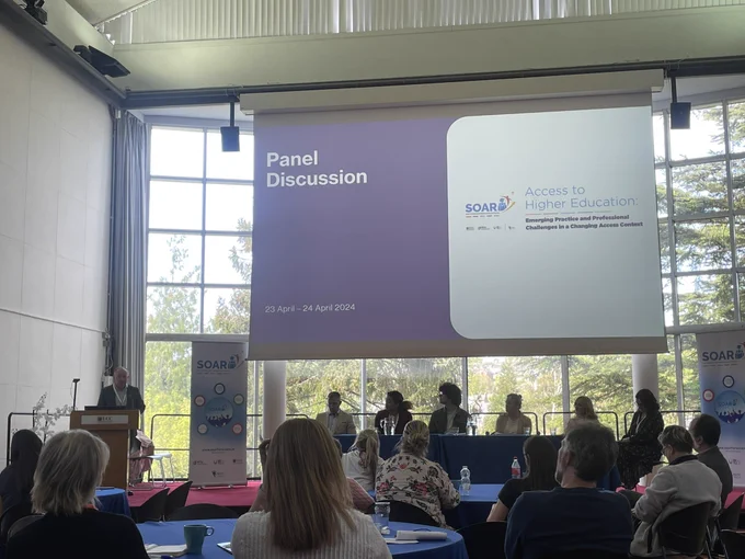 Man at podium in front of large window and large screen to his right.  Panel of 6 people sitting to his right under the screen