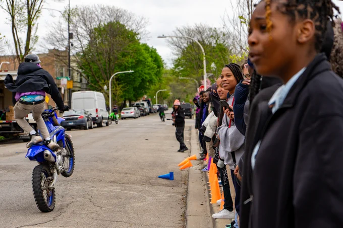 Children smile as they watch a motorcyclists pop a wheelie while standing on the seat of his bike. 