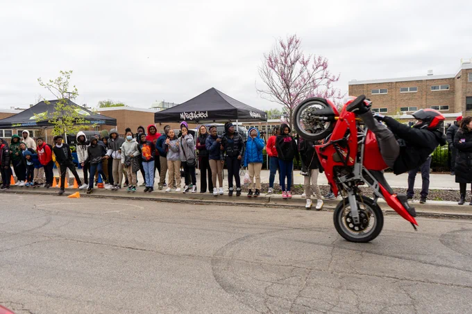 Motorcyclist in the foreground on the street popping a wheelie with his legs spread wide. Middle school aged children stand in the background watching. 