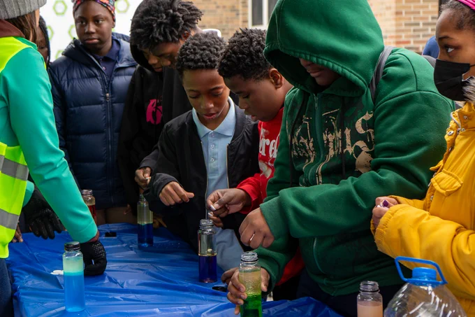 Students crowded around a table drop white tablets into jars of colorful liquid. 