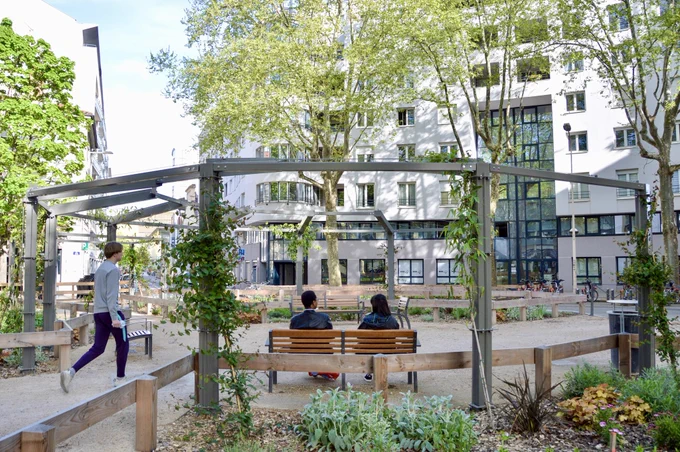 People walk, cycle, and sit in a “Super Island” in Lyon— an intersection transformed into a public space by introducing traffic calming, trees and plants, street furniture, and bike parking.