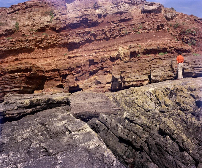 SE coast, Sully Island. Looking N. Triassic - Carboniferous unconformity, Sully Island. Red Triassic strata in the background with a small fault in top right of photograph. Carboniferous 'Black Rock Dolomite' in the foreground dips north-east at 45 degrees The 'planed-off' surface represents the exhumed Triassic land surface. Small pebbles and flakes of Carboniferous Limestone in calcareous sandstone matrix cover the limestone on the left, the figure on the right (height 1.7 m.) is standing on a surface of bare limestone. The basal Triassic Clastic Unit can be seen to thicken towards the left.
