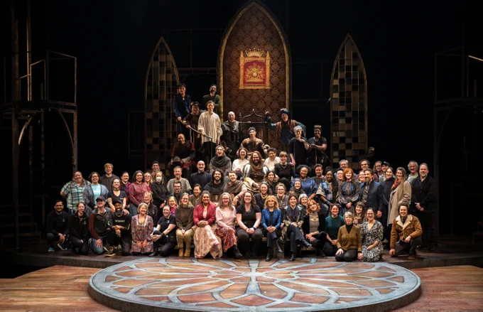 The cast, crew and creative team of HENRY V sit stacked on the stage in front of the set, smiling for the photo.