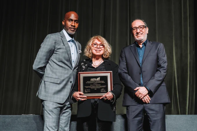 Three people pose for a photo and smile at the camera. The person in the middle is holding an award plaque.