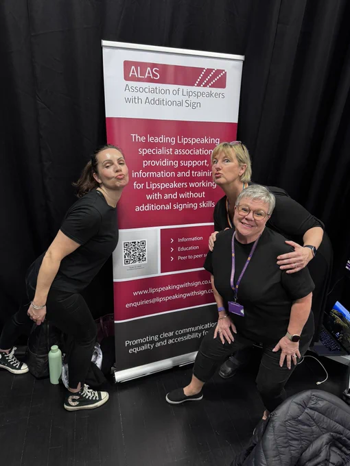 Three women posing for a picture, with a banner, reading ‘The Association of Lipspeakers with Additional Sign’.