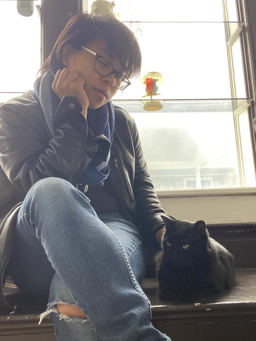 Sean sitting by a window, legs crossed. A black cat named Peep sits beside him.