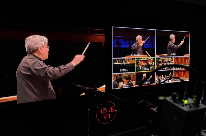 A backstage view of a television gallery showing two screens with images of an orchestra playing.