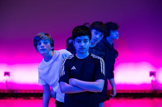 6 teenage boys are in a line. The boy at the front has a serious expression and his arms are folded. Those peeking out behind him are looking in different directions with milder expressions. They are bathed in blue light but the wall behind them is lit up pink.