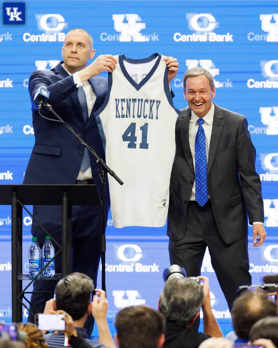 Mark Pope holding up his 1996 Kentucky jersey, Mitch Barnhart standing next to him smiling. 