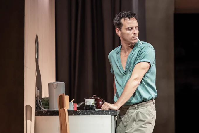 Andrew Scott wears a blue silk shirt and stands at a kitchen work surface. He holds a cigarette in his mouth.
