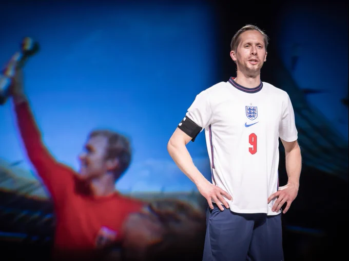 A man in an England white football shirt and with a black band around his arm stands with his hands on his hips.