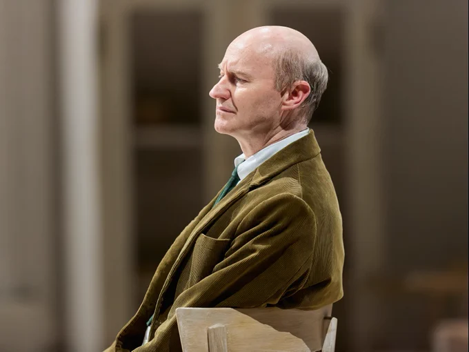 A man in an olive green corduroy suit jacket and green tie sits on a wooden chair and looks deep in thought.