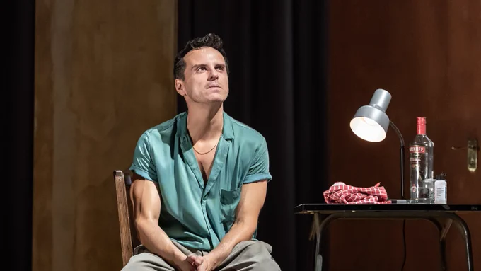 A man in a blue shirt and brown trousers sits in a chair and looks deep in thought. Next to him on a table is a vodka bottle and small lamp.