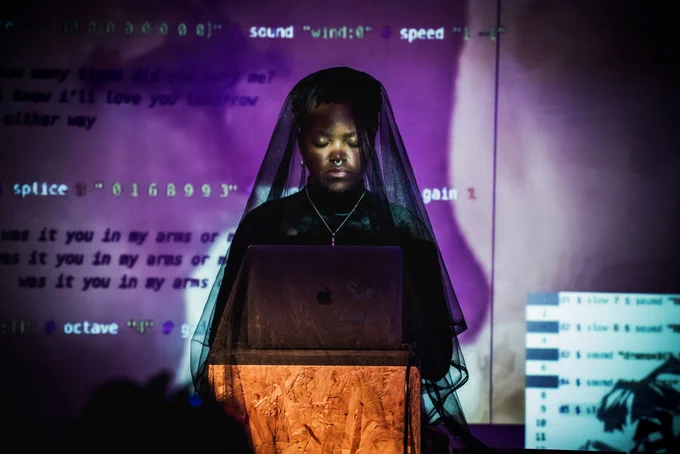 Performer wearing a black veil which covers themselves and a laptop. They are illuminated by a purple projection of computer code.