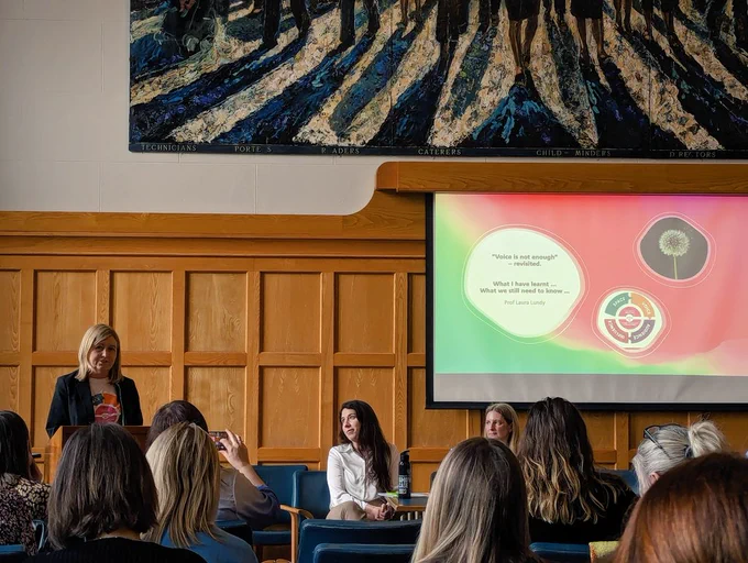 Professor Laura Lundy, a white female, presents to a busy room with people sitting down listening. On the screen are bright colours with a picture of a dandelion and the name of the event. 