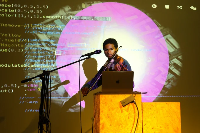 Performer playing violin into a microphone behind a wooden platform with a laptop on it. They are illuminated by a large projection of computer code.