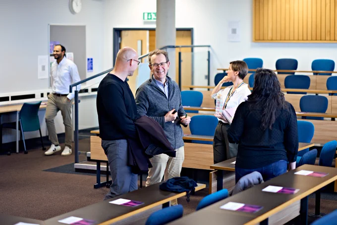 Group of researchers stood talking and smiling in a lecture theatre