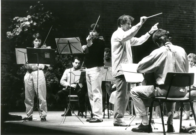 A group of musicians on stage, cellists sitting  and violinists standing, with a conductor amidst them.  All are in casual clothes, as this is a rehearsal rather than performance.