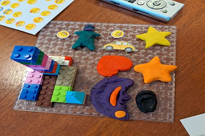 Photograph of a clear a lego base with some lego built up on it and a person, a star and an ear in playdough o it with a sticker of a car and some smiley faces. 