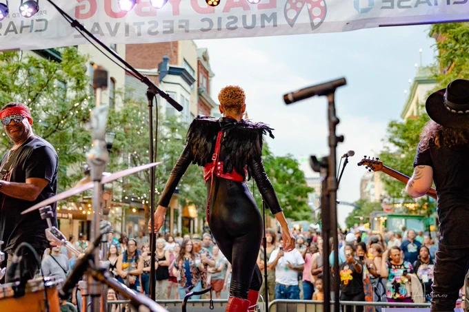 A musician with a mohawk and red outfit stands on stage facing a crowd, with band members beside her and banners overhead.