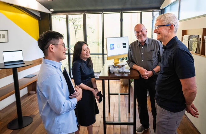 Tim and three developers stand beside a tall table chatting. Apple Vision Pro and iMac are on the table. MacBook Air is on a circular table on the left against a white and yellow wall.