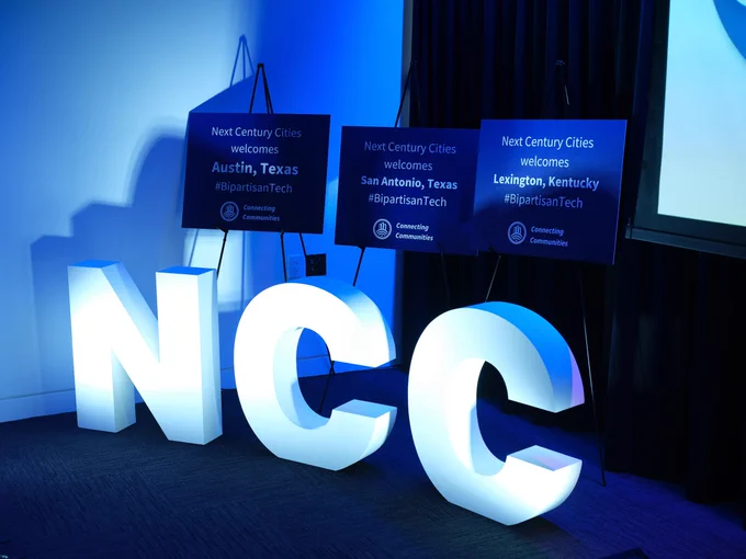 Photo of Next Century Cities' logo in front of signs welcoming guests from various cities to the Bipartisan Tech Conference.
