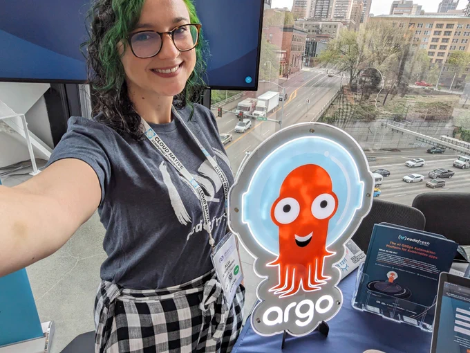 Selfie of me, a white woman in her thirties with dark brown and green curly hair in a Codefresh tshirt next to a light up Argo sign 