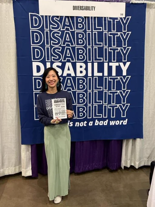 Tiffany is holding The Anti-Ableist Manifesto art on a postcard standing in front of a "Disability Is Not A Bad Word" photo backdrop that is also a shower curtain!