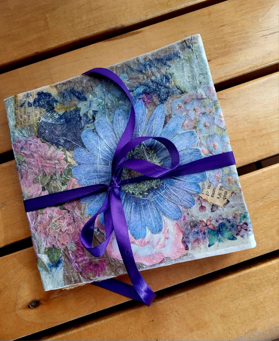A small handmade journal with blue and pink flowers surrounded with a purple bow.