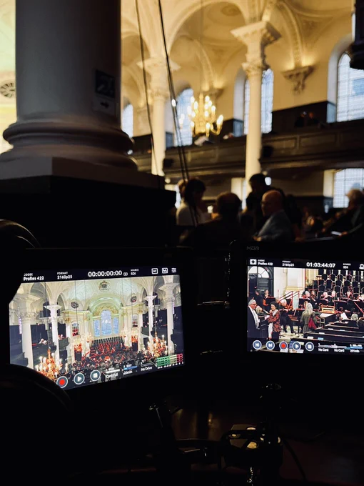 Behind the scenes at St Martin-in-the-Fields in London, with audience milling in the background. Two small video screens show camera shots of the church’s brightly lit interior.