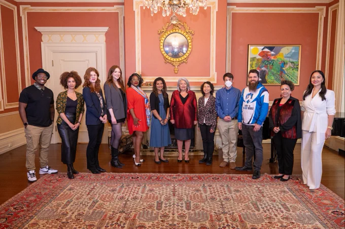 Governor General Mary Simon is standing among a group of 12 people. They are posing for a photo. There is a large rug at their feet. 